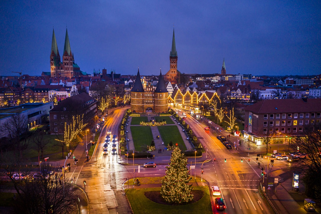 Abendliche Stadtansicht von Lübeck mit beleuchtetem Holstentor und Weihnachtsbaum und den Kirchtürmen im Hintergrund.